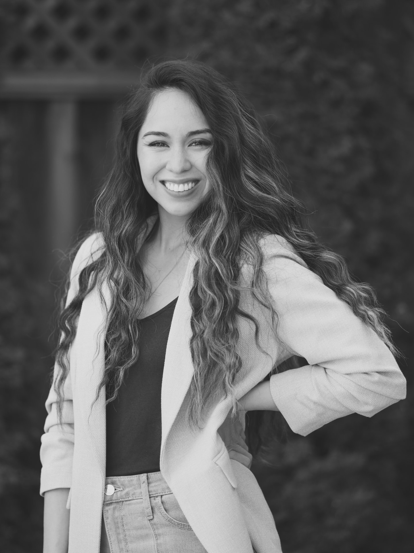 Linnea Locsin, coach at The Voice Collective Co., photographed in black and white outdoors in front of a hedge, smiling with her hand on her hip