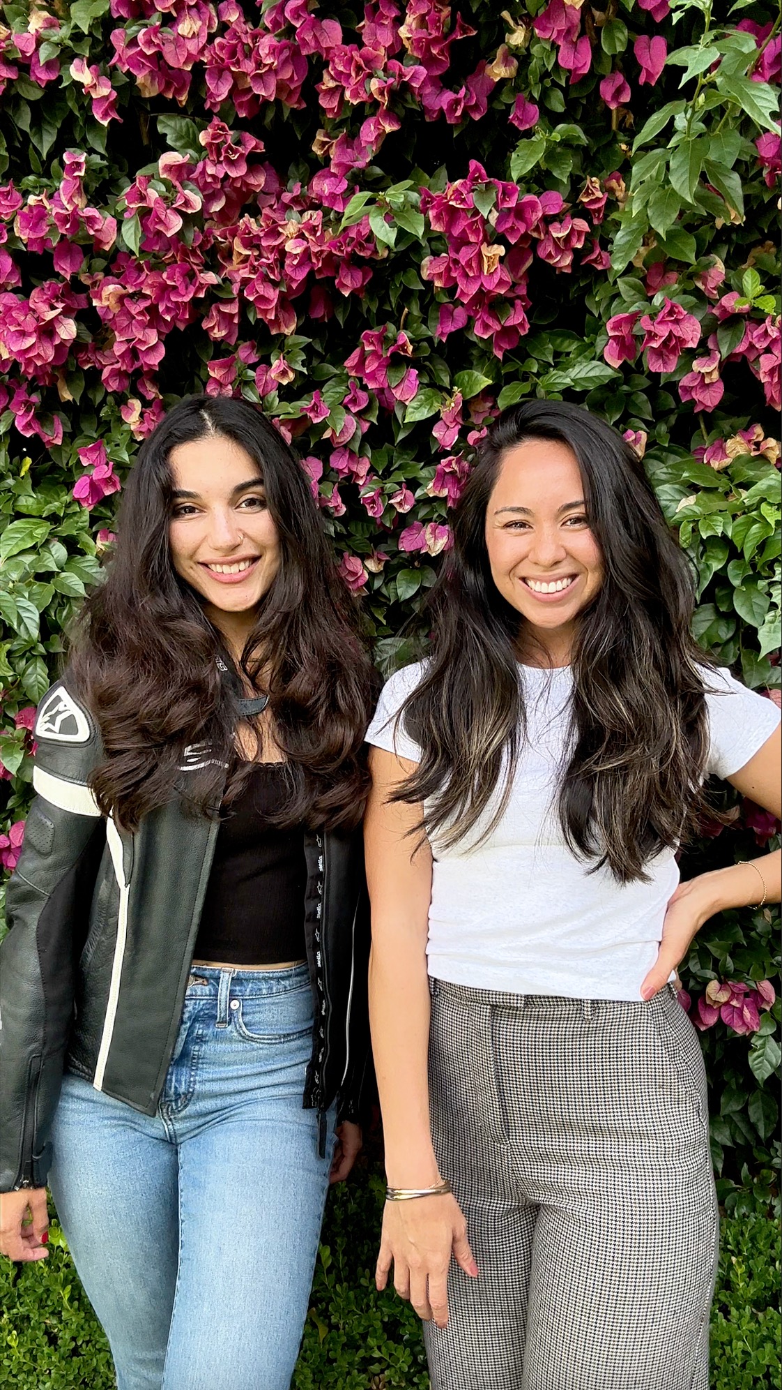 Two members of the collective smiling side by side in front of a wall of bougainvillea