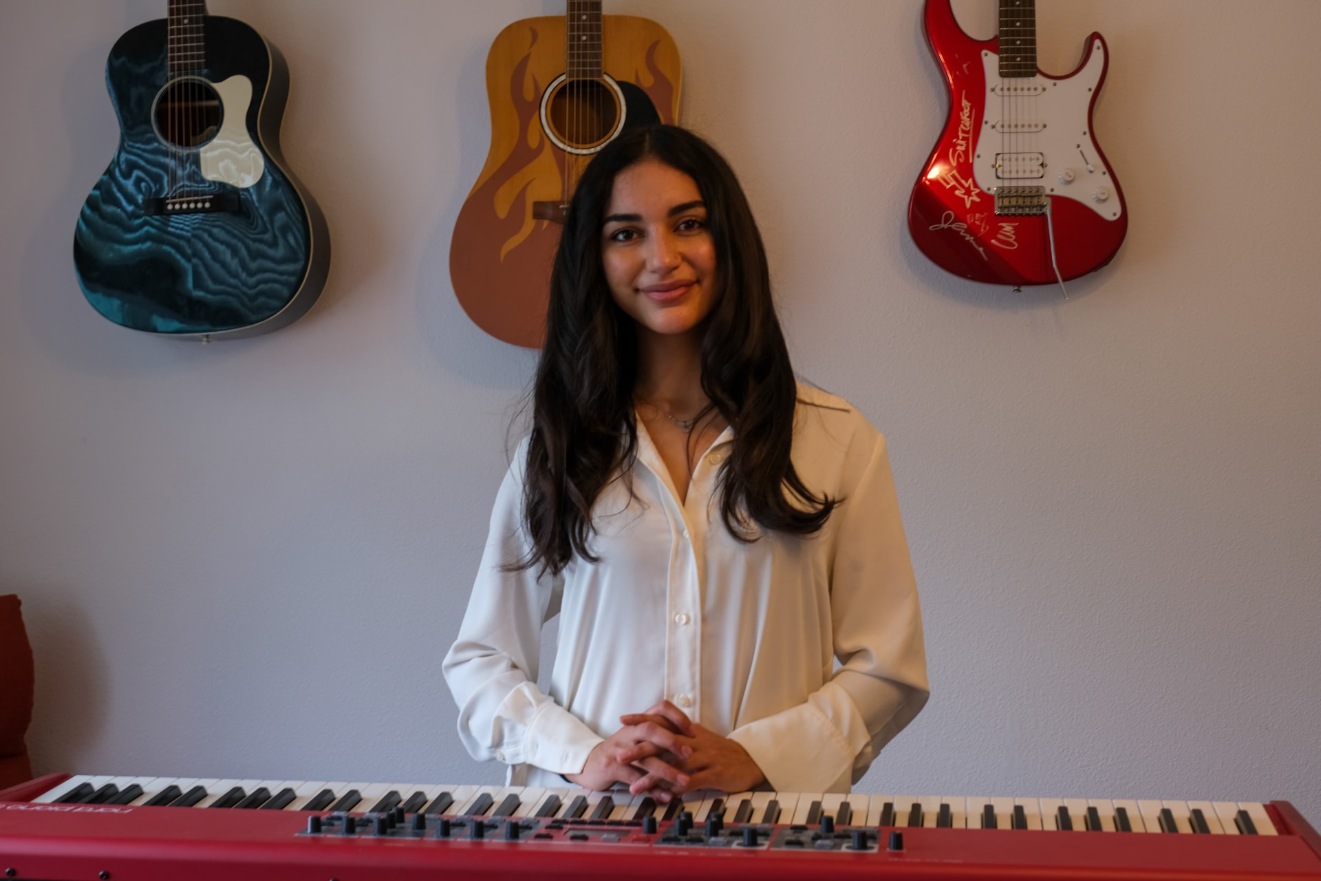 A vocalist standing behind a keyboard with guitars hanging on the wall behind her